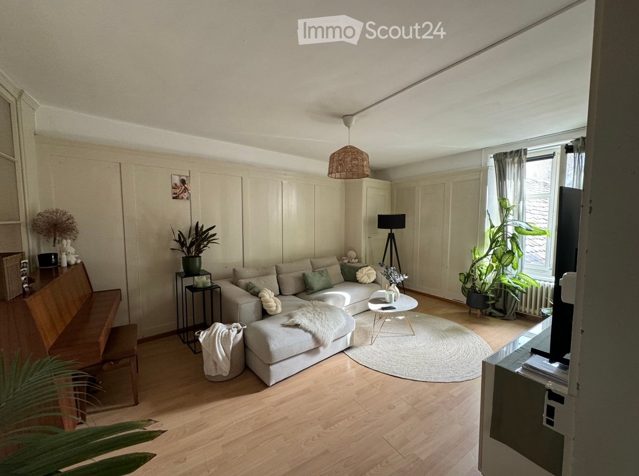 Modern living room with light-colored sofa, coffee table, bench, floor lamp, and plants. The room features wooden flooring and large windows with curtains.