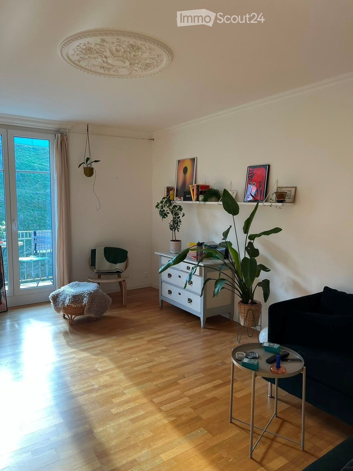 Modern living room with wooden flooring, large windows, balcony access, sofa, coffee table, small stool, chair, table, and plants.