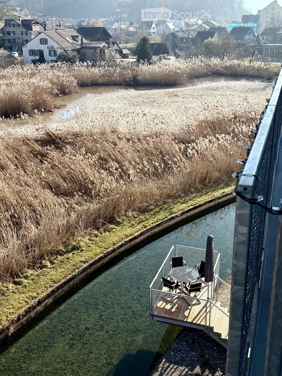 Balcony overlooking a river with a table, chairs, and an umbrella, surrounded by reeds and a small town in the background.