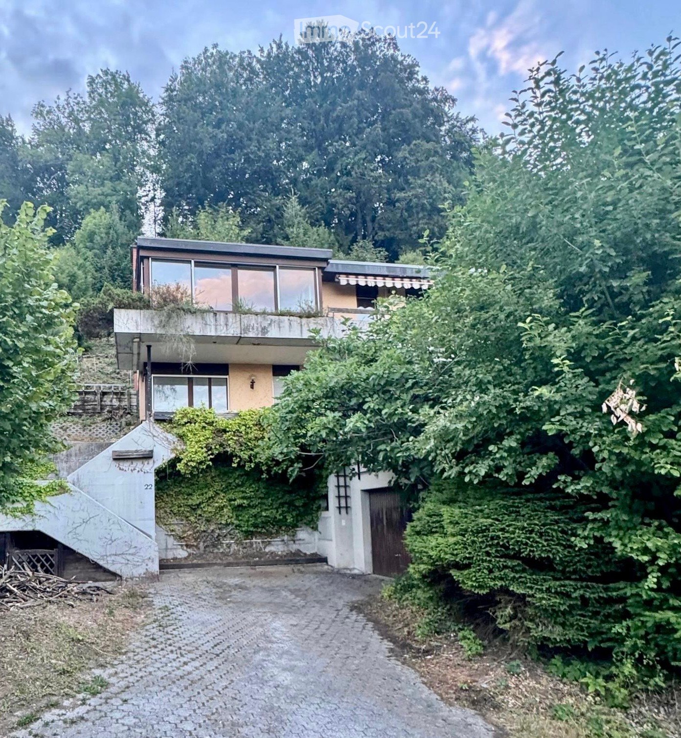 Modern two-story house with large windows, flat roof, and surrounding greenery. Features a raised ground floor and a covered entrance area.