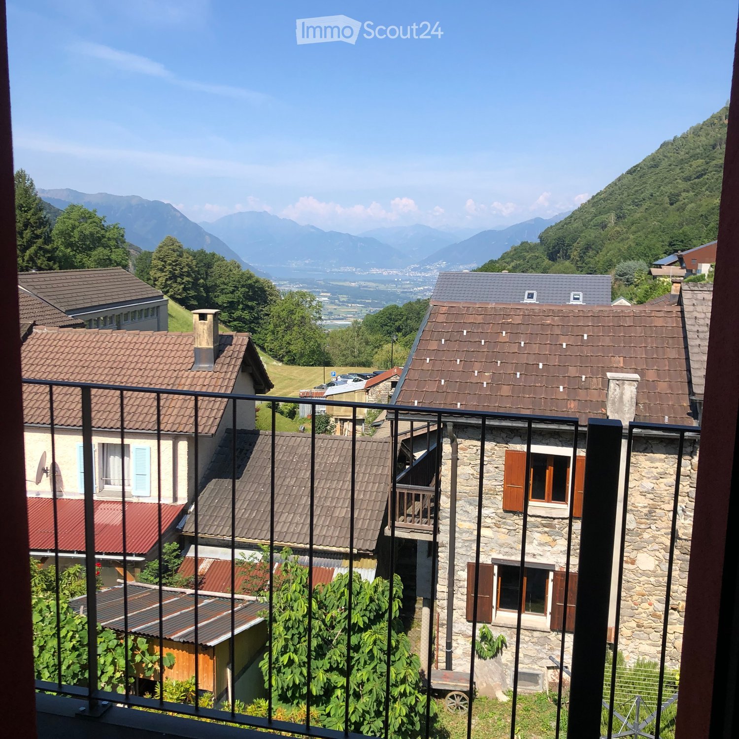 Balcony overlooking a village with stone houses, tiled roofs, and distant mountains under a clear blue sky.