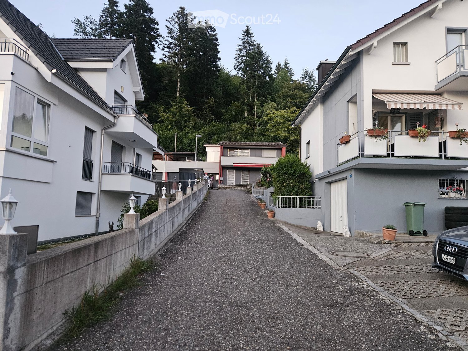 Residential street with modern two-story houses, balconies, and greenery in the background.