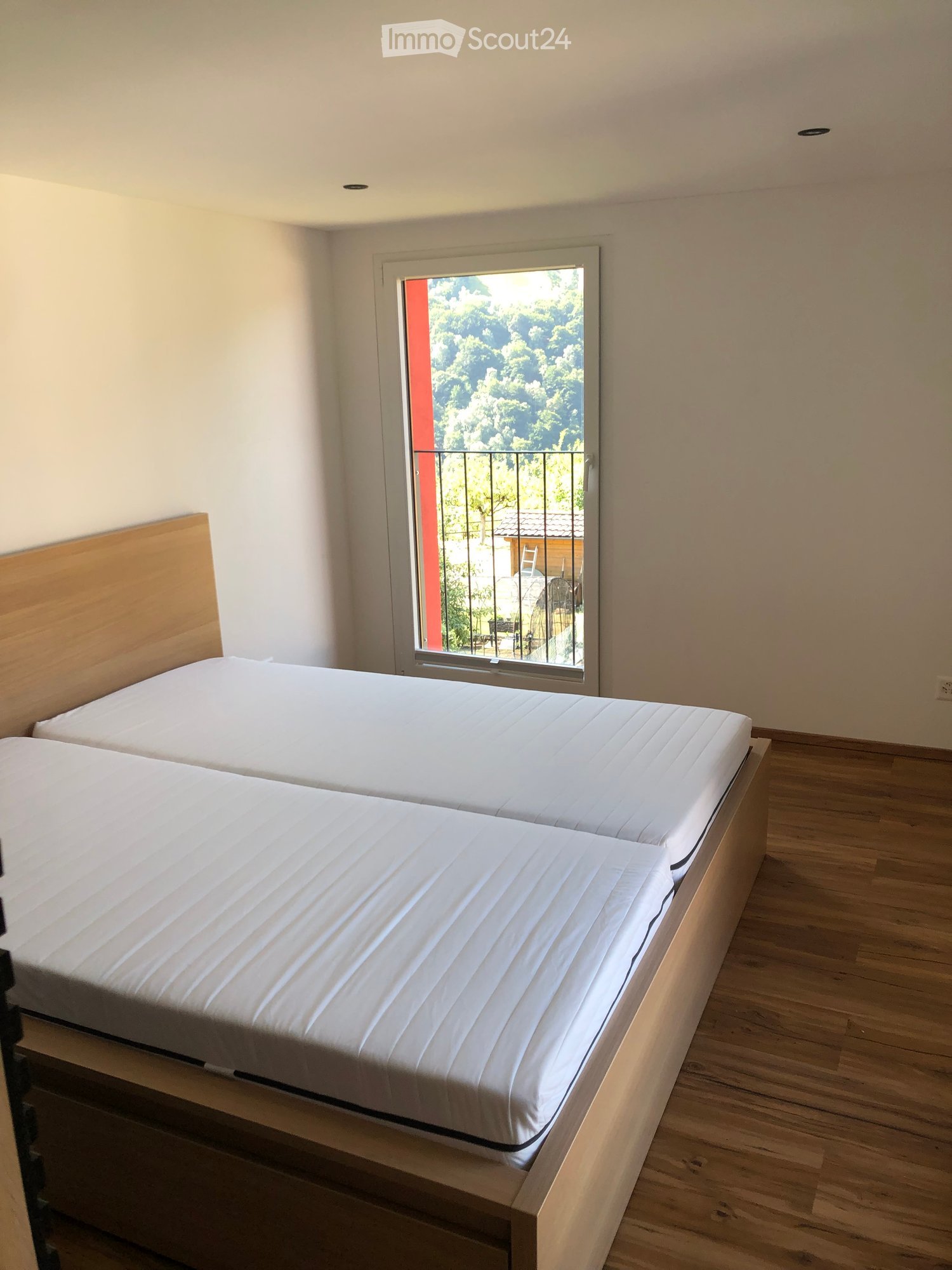 Bright bedroom with two single beds, wooden headboards, and a large window with a view of greenery and a balcony.