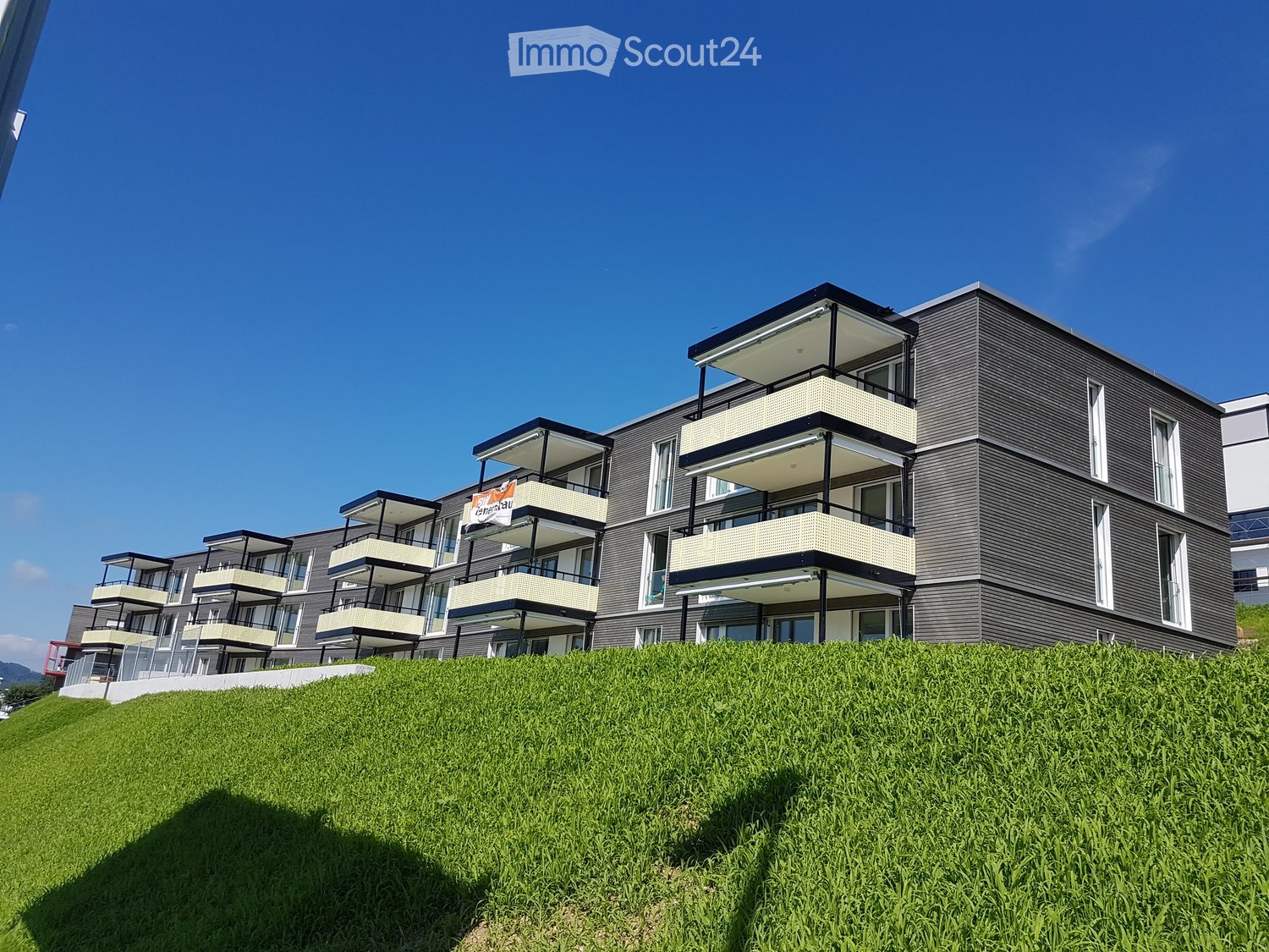 Modern multi-story residential building with balconies and large windows, situated on a grassy slope.