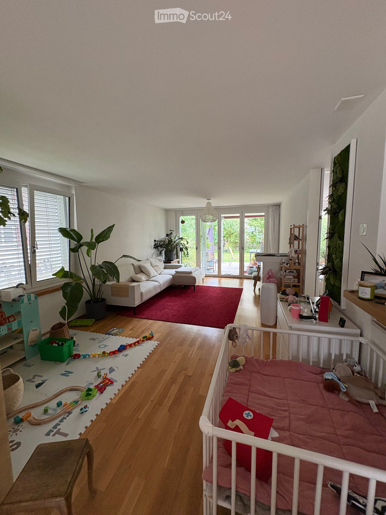 A well-lit living room with a white sofa, ottoman, and a red rug. A crib with a pink blanket and toys is visible, along with a play area featuring a toy train and cars. There are plants, a shelf, and a table with a laptop and cup.