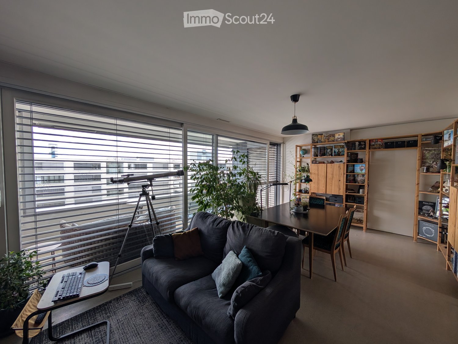 Modern living room with large windows, grey sofa, laptop on side table, dining table with chairs, bookshelf, and potted plants.