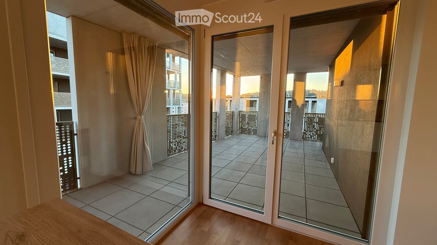 View of a tiled balcony area accessible through large glass sliding doors from an interior space with wooden flooring.