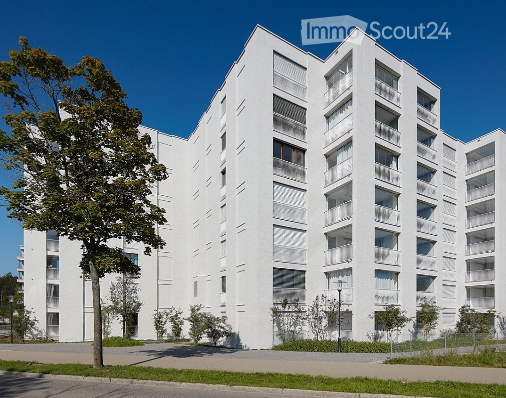 Modern white apartment building with multiple balconies and large windows, surrounded by greenery and a paved area.