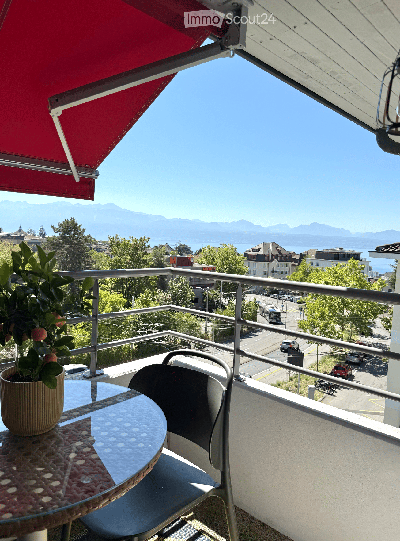 Balcony with a view of a cityscape and mountains, featuring a table, chair, and potted plant.