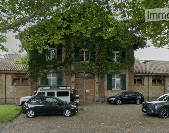 A two-story brick building with green shutters and a large wooden door, surrounded by trees. Several cars are parked in front of the building.