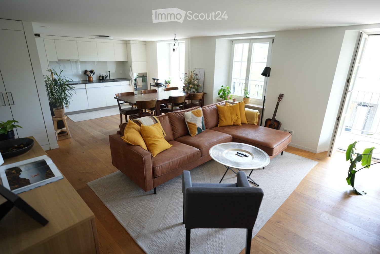 Modern living room with a brown leather sofa, yellow cushions, a round coffee table, a grey armchair, a dining area with a table and chairs, a kitchen area with white cabinets, and various plants and decor.
