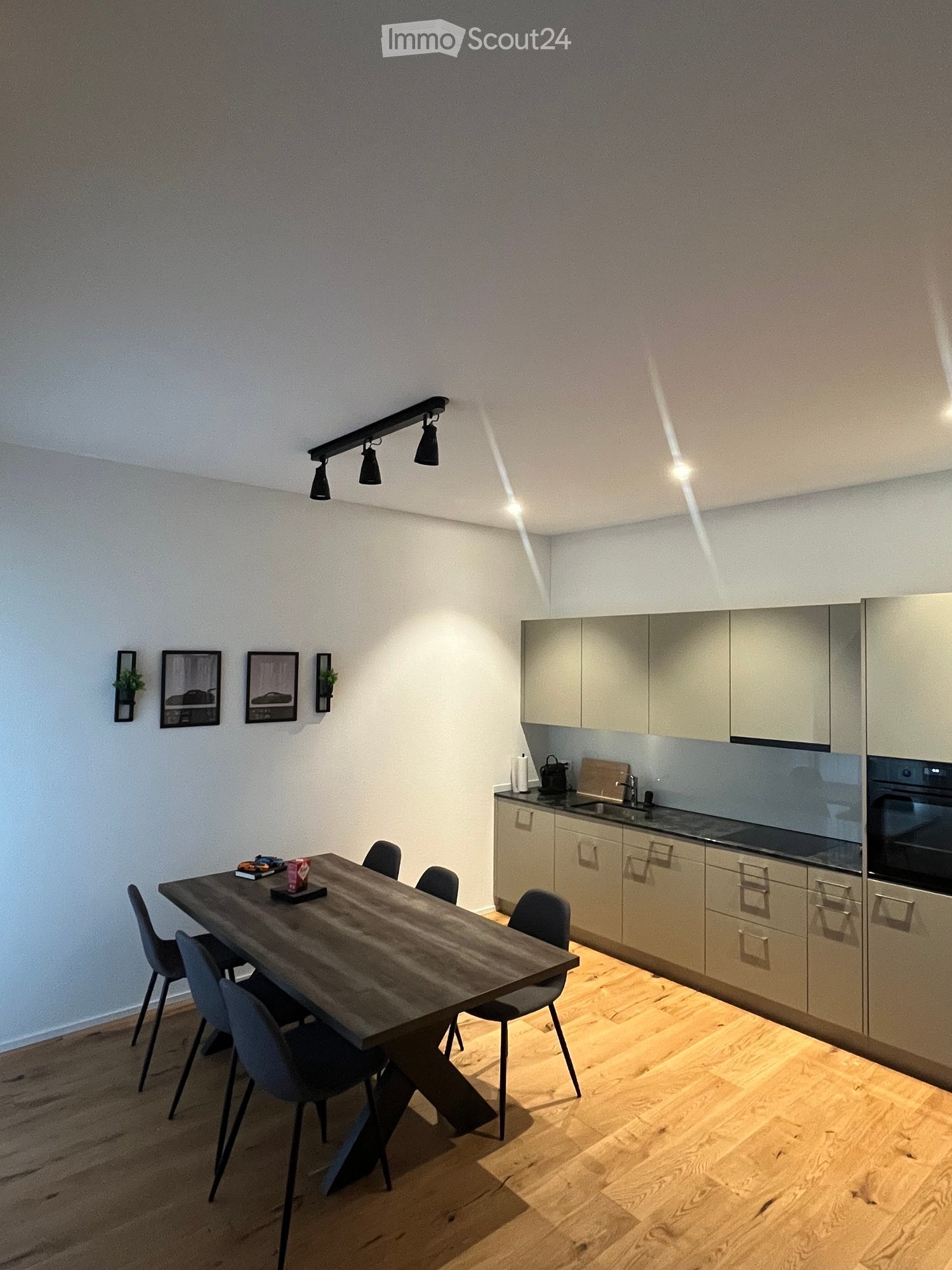 Modern kitchen with light wood cabinets, stainless steel appliances, a wooden dining table with chairs, and wall-mounted artwork.