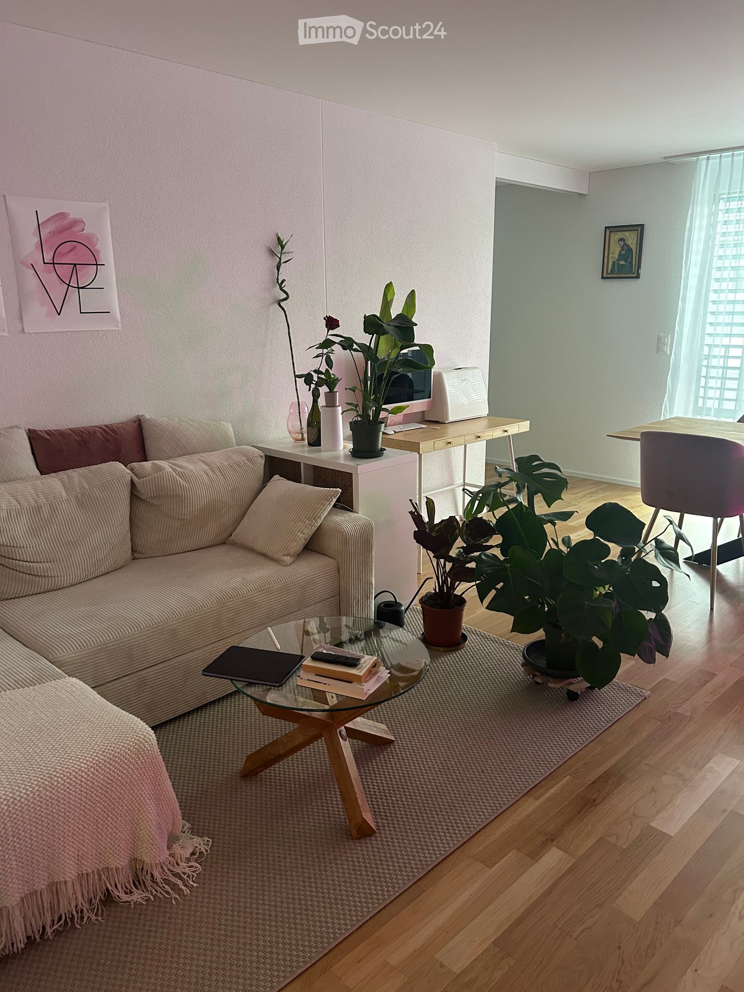 A modern living room with a beige sofa, glass coffee table, wooden floor, and several potted plants. There is a small desk with a laptop and a chair in the corner, along with a blanket draped over the sofa.