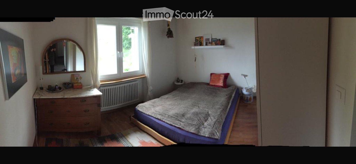Cozy bedroom with a double bed, dresser, mirror, and shelves. The room features wooden flooring and large windows with white curtains.