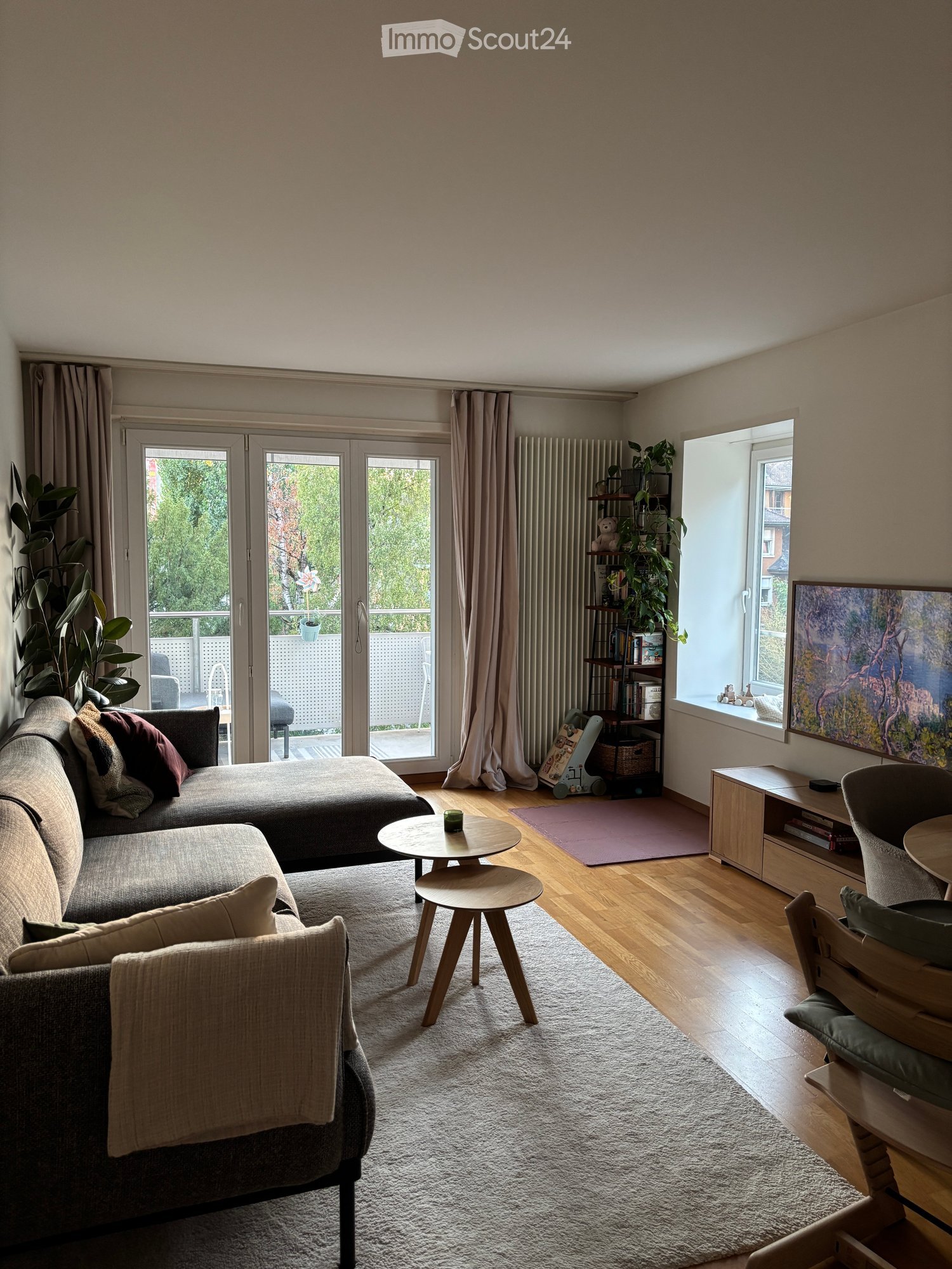 Modern living room with a grey sectional sofa, wooden coffee tables, a white shag rug, and large windows leading to a balcony.