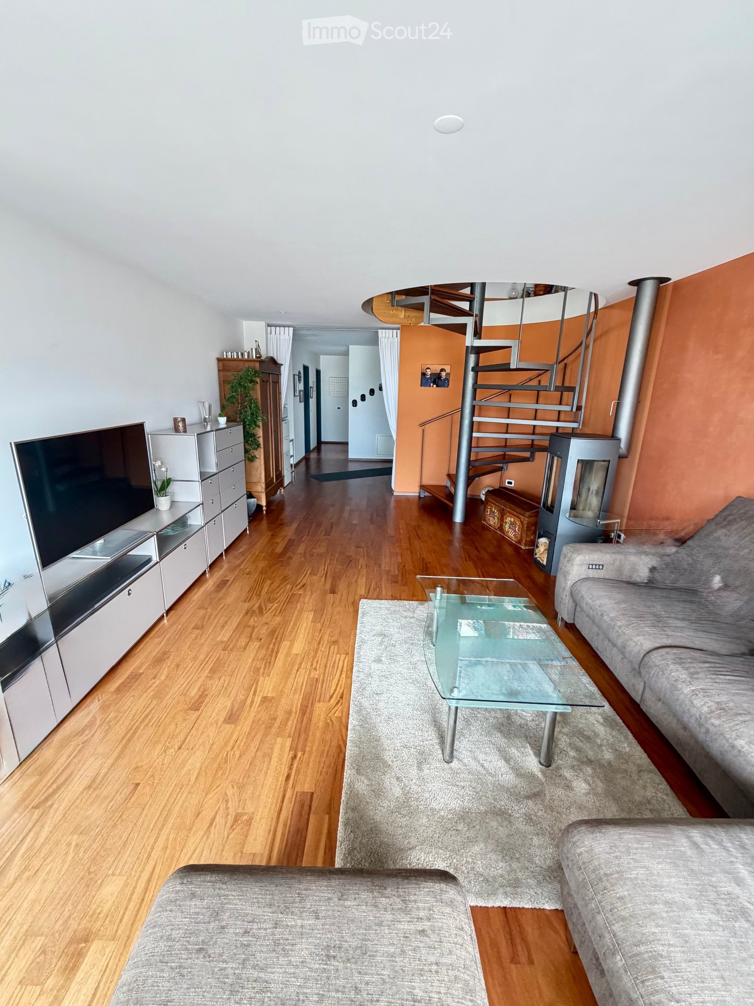 Modern living room with hardwood flooring, grey sofa, glass coffee table, flat-screen TV, and a spiral staircase leading to an upper level.