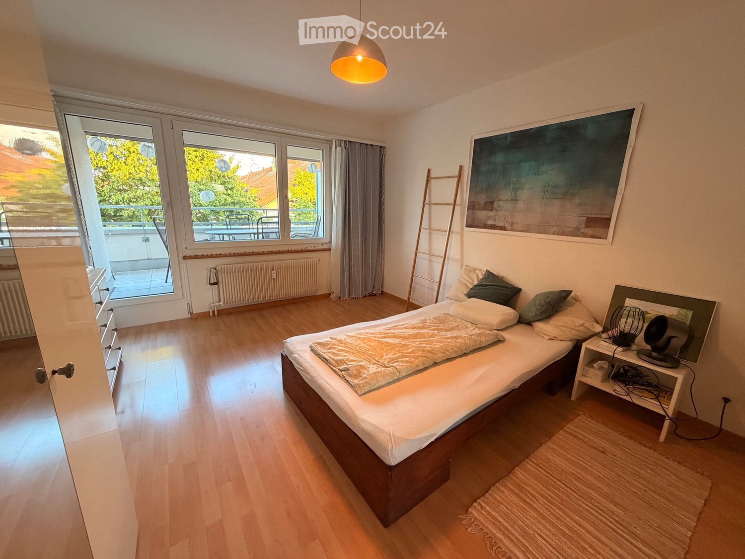 Cozy bedroom with wooden flooring, a double bed with pillows and a blanket, a nightstand with a lamp, a mirror, and a ladder leaning against the wall. Large windows offer a view of a balcony with outdoor furniture.