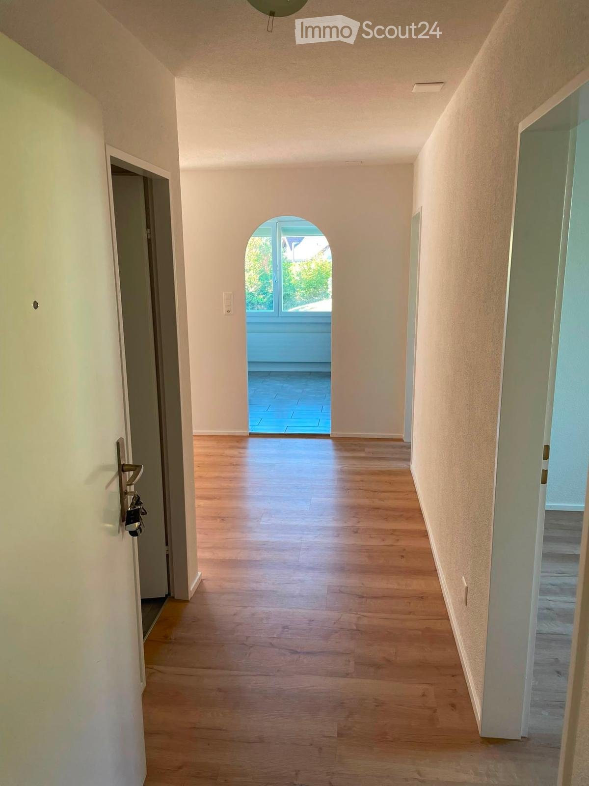 Empty hallway with wooden flooring, white walls, and a small arched window at the end leading to a blue-tiled area.