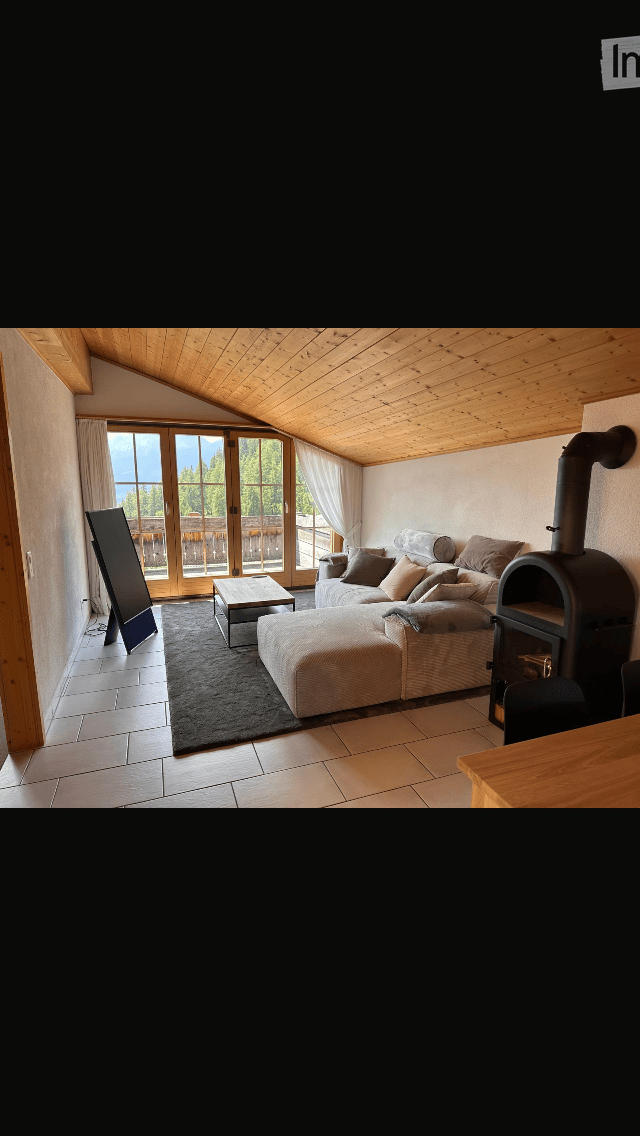 Bright living room with wooden ceiling, large windows offering a scenic view, a sofa, coffee table, ottoman, and a fireplace.