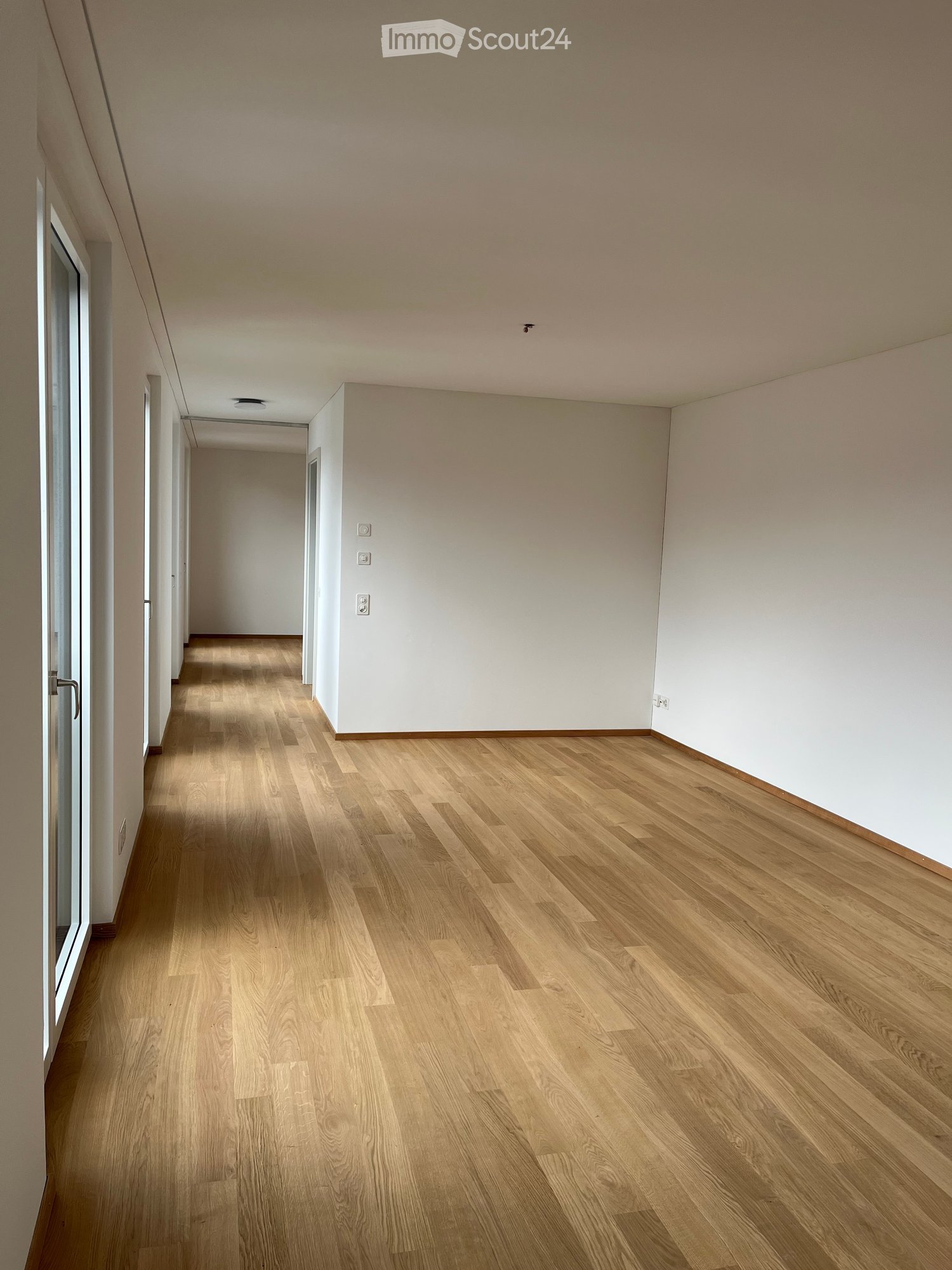 Empty hallway with light wooden flooring, white walls, and recessed lighting. Features a door on the left and electrical outlets on the right wall.