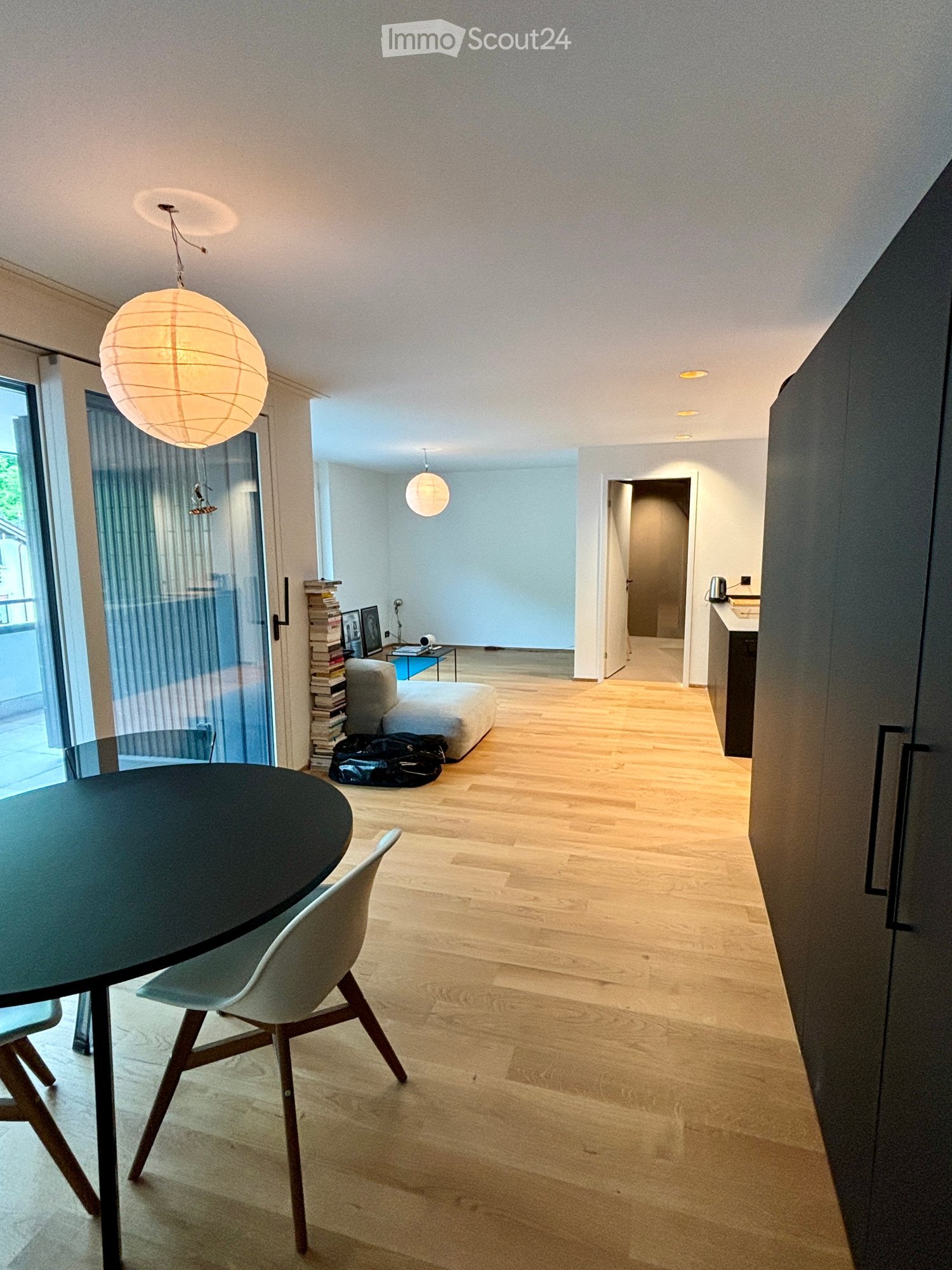 Modern, open-plan living and dining area with wooden flooring, large pendant lights, a round dining table with chairs, a sofa, a stack of books, and a kitchen area visible in the background.