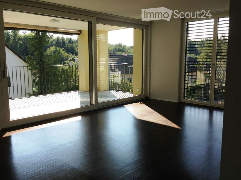 Empty room with dark wooden flooring, large sliding glass doors leading to a balcony with metal railings, and a view of greenery and neighboring buildings.