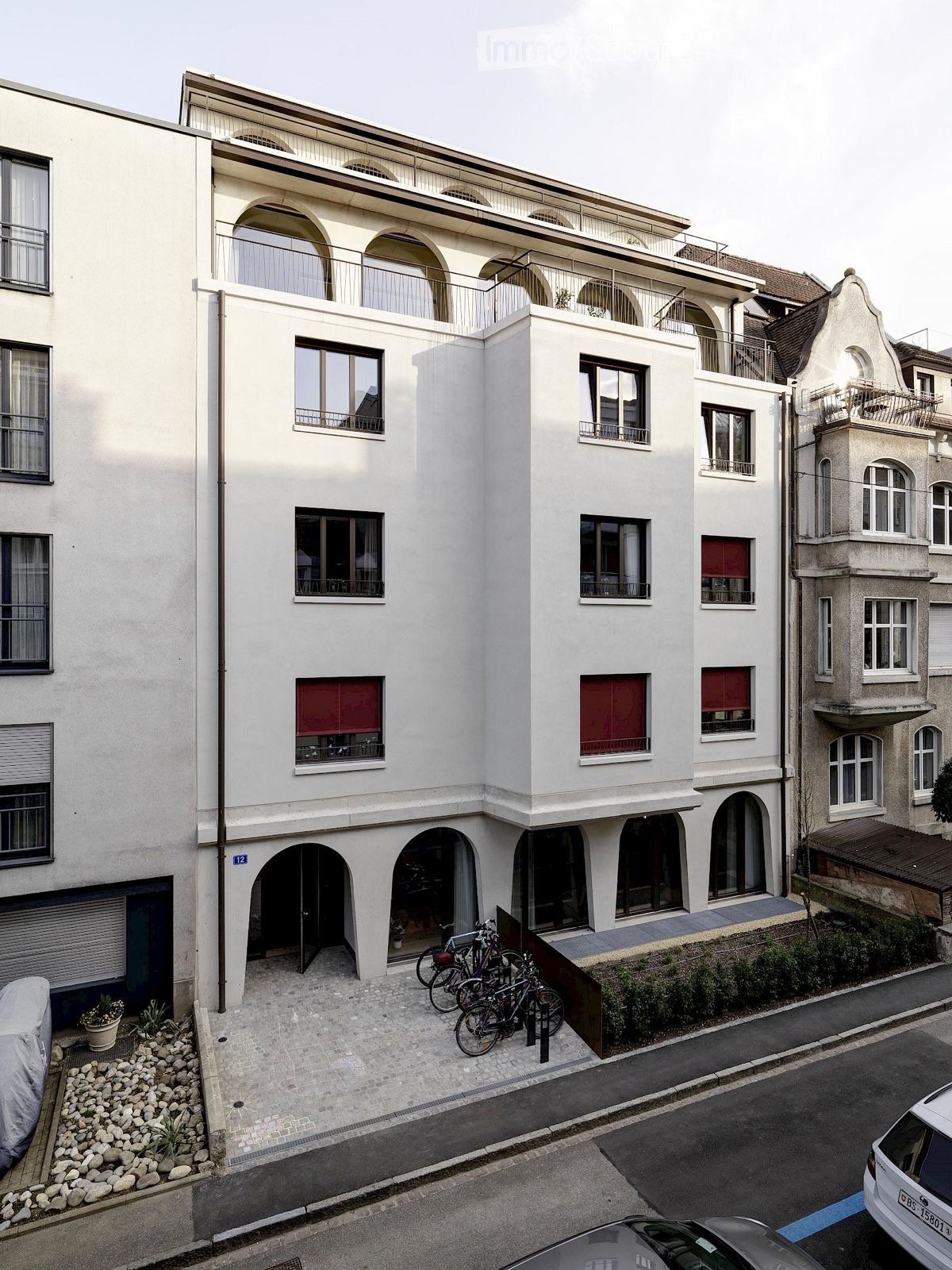 Modern multi-story building with white facade, arched windows, and balconies. Bicycles parked in front, small garden area with potted plants, and a covered car parked nearby.