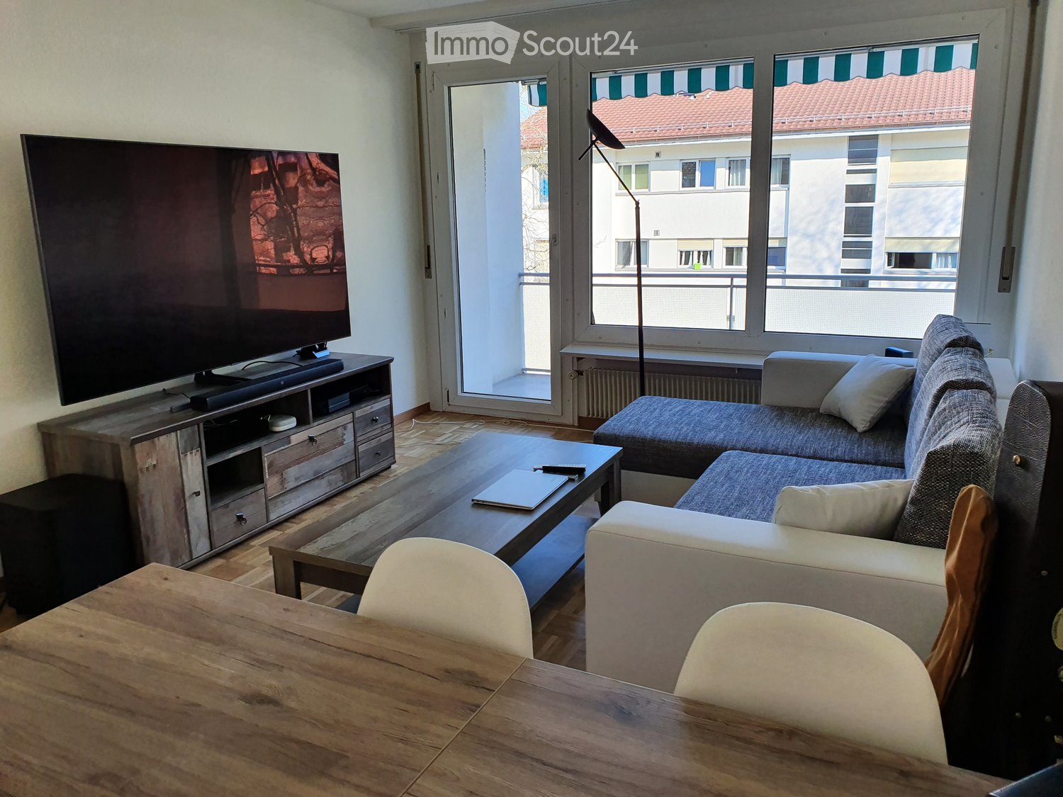 Modern living room with a grey sectional sofa, coffee table, TV stand, and dining table with chairs. Large windows and a sliding glass door lead to a balcony.