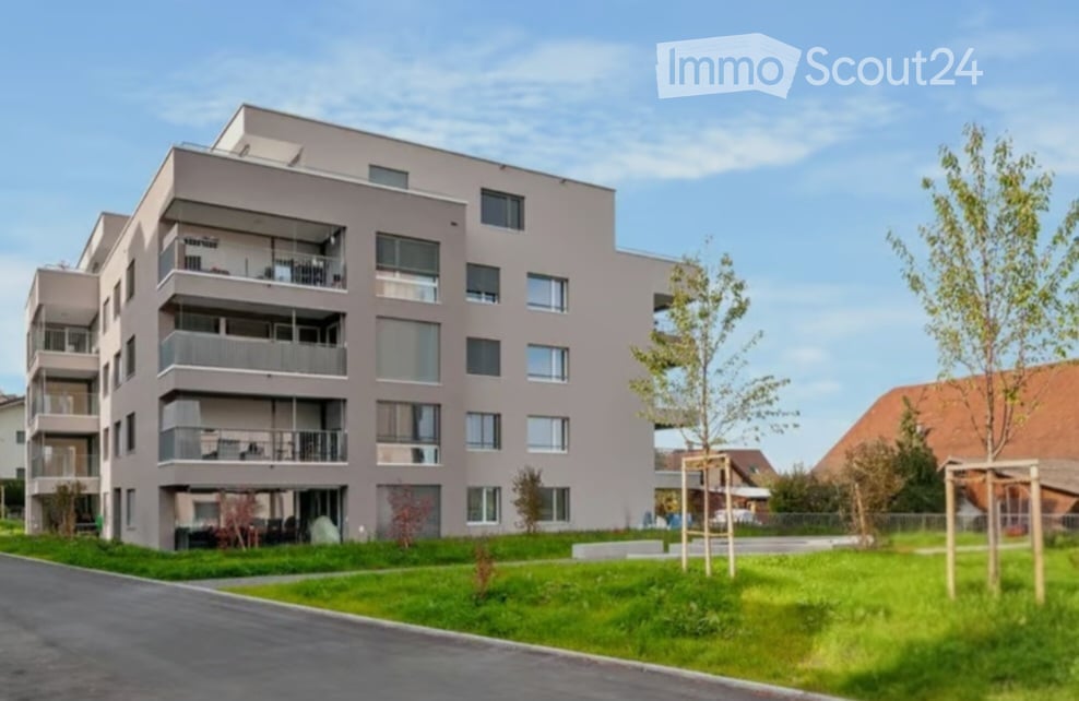 Modern multi-story apartment building with balconies, large windows, and a landscaped green area in front.