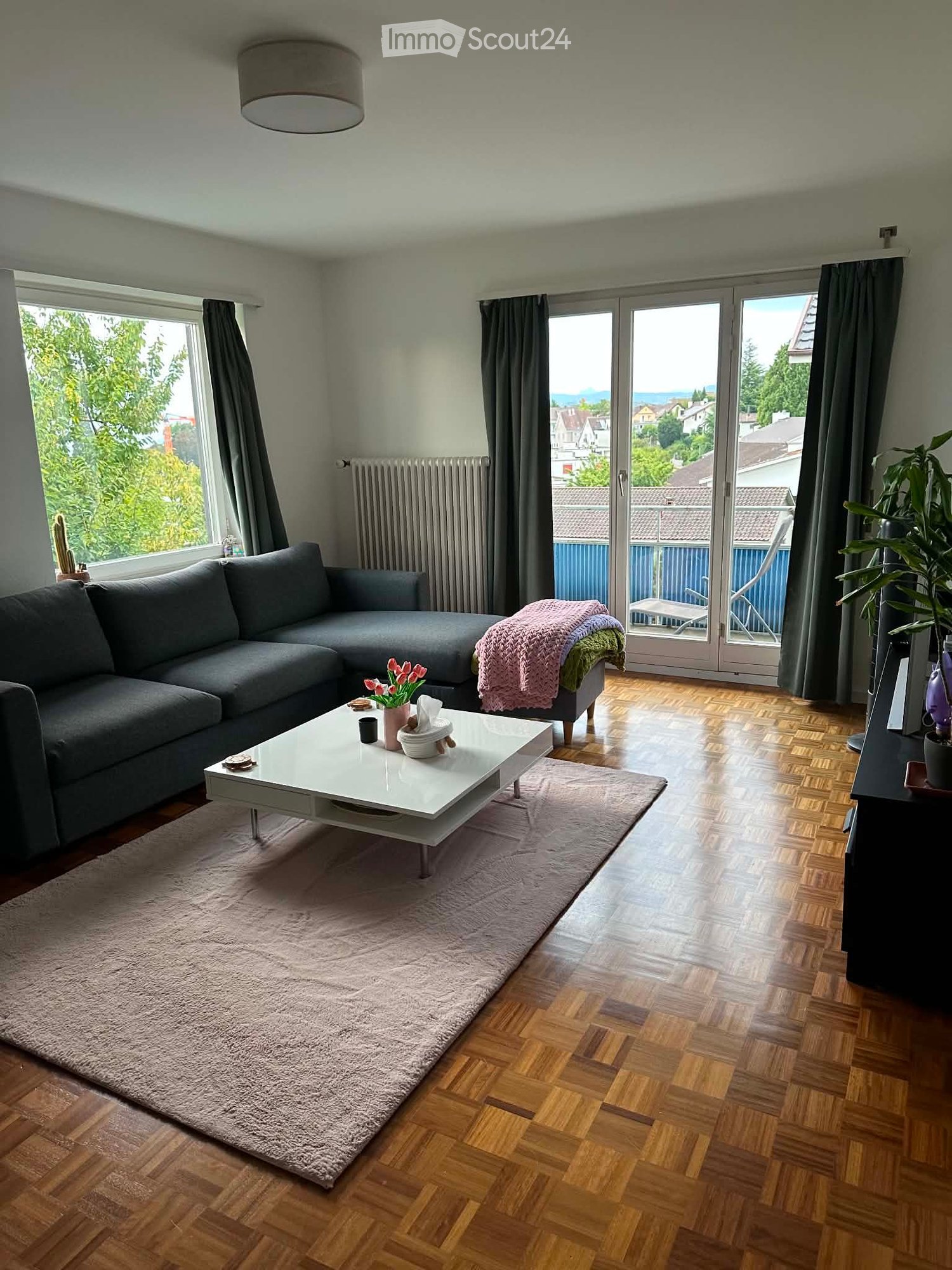 Modern living room with wooden flooring, large windows, grey sectional sofa, white coffee table, and a view of the city.