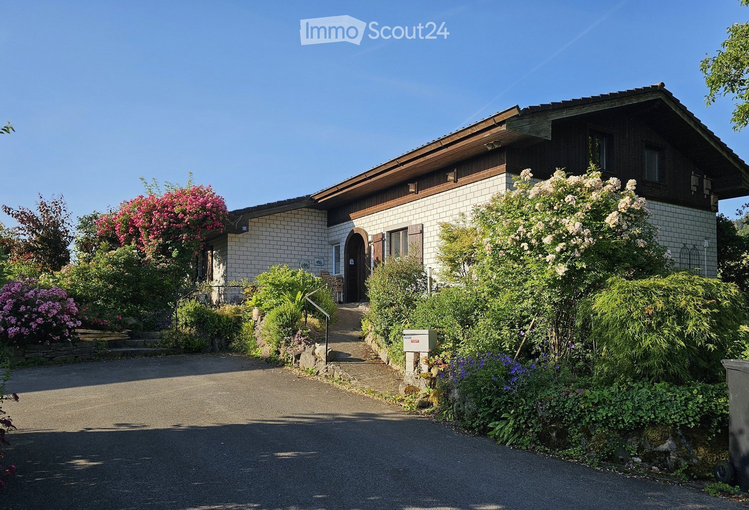 Two-story house with a tiled roof, white brick facade, and a well-maintained garden with colorful flowers and shrubs.