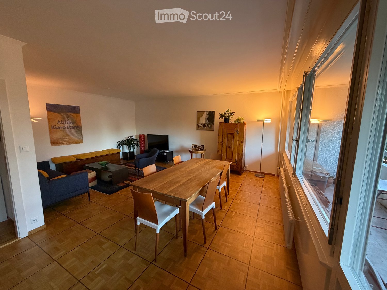 Bright, open living and dining area with wooden flooring, large windows, a sofa, coffee table, dining table with chairs, TV, and decorative items.