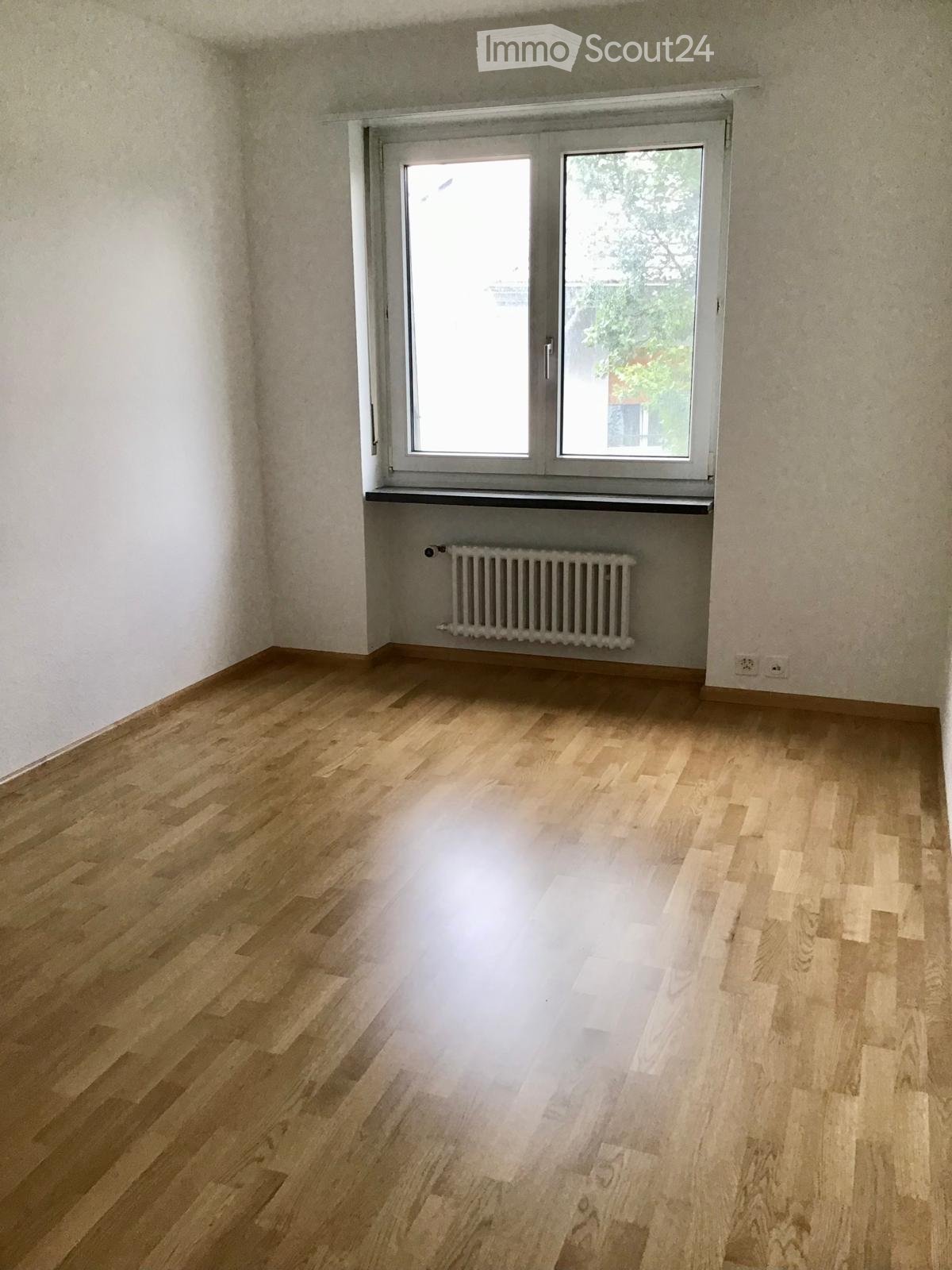 Empty room with light wooden flooring, white walls, and a large window with a radiator below it.