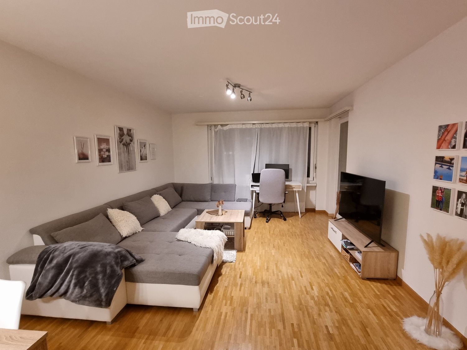 Modern living room with a grey sectional sofa, wooden coffee table, desk with chair, TV stand, and decorative vases.