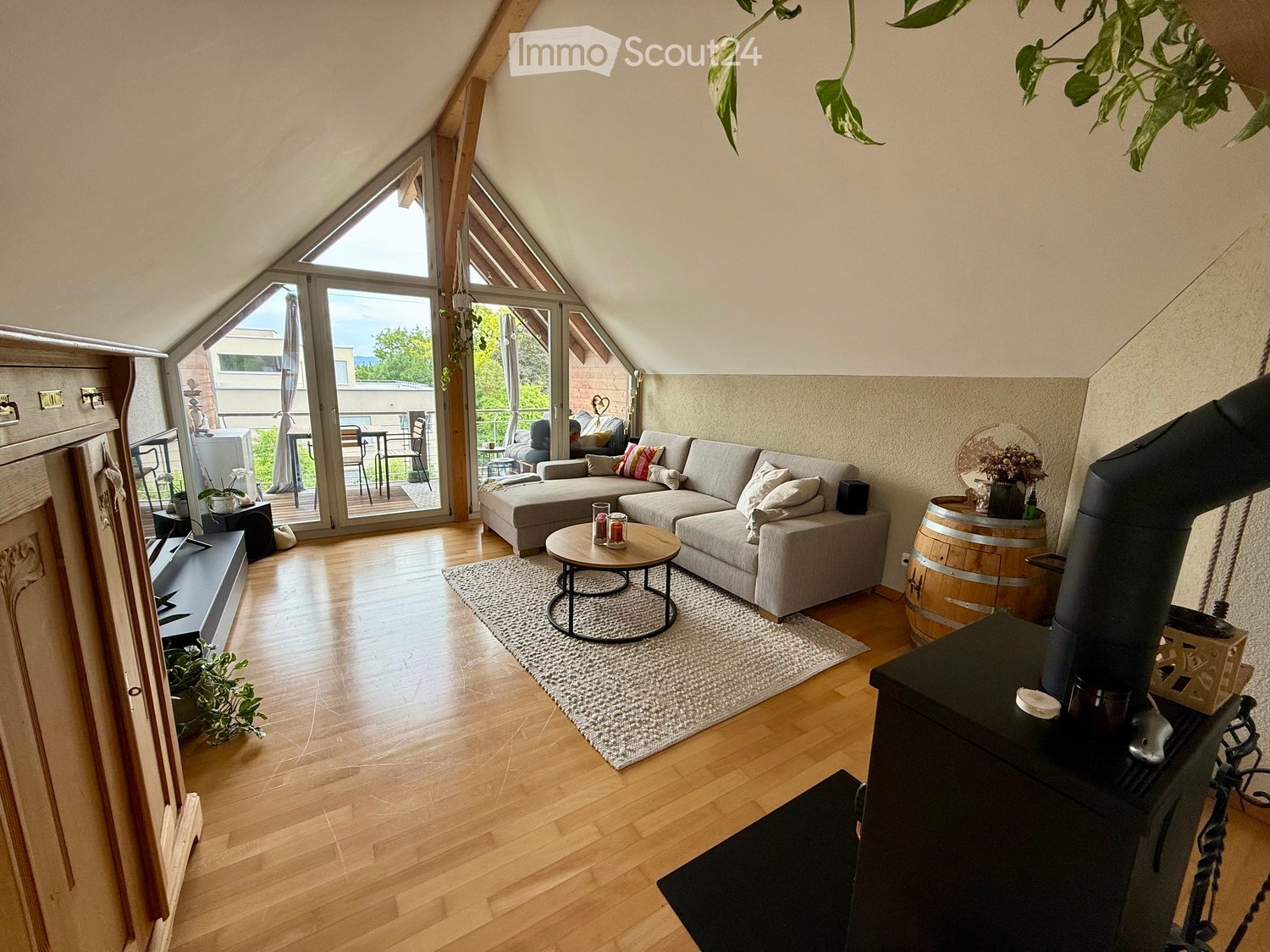 Bright living room with wooden floors, large windows, a grey sectional sofa, coffee table, wood stove, and a view to a balcony area with outdoor furniture.