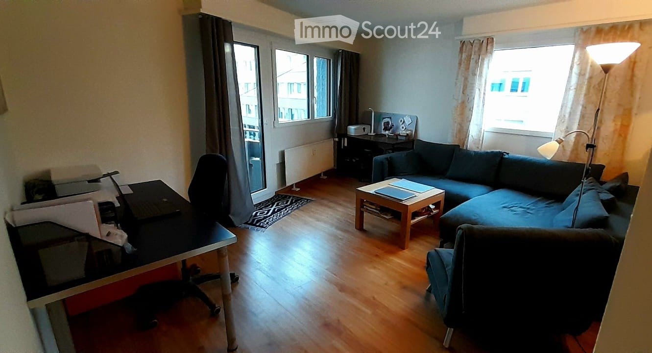 Modern living room with wooden flooring, large windows, a sectional sofa, coffee table, desk with laptop, and a floor lamp.