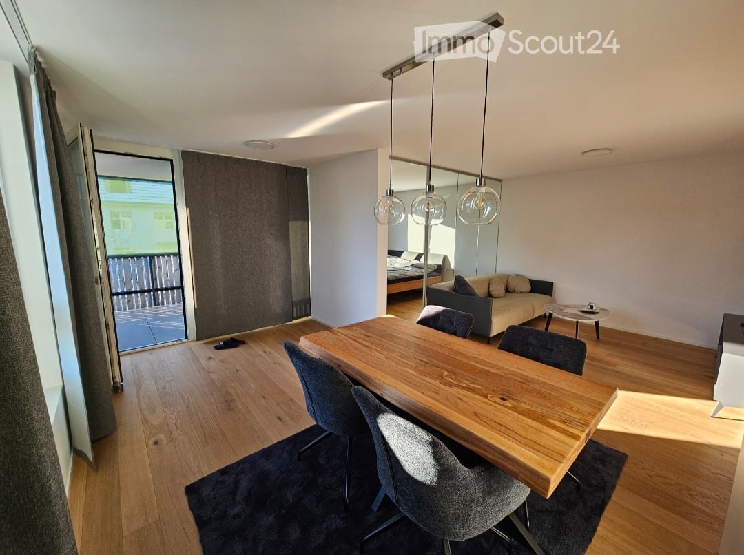 Open-plan living and dining area with wooden flooring, a large wooden dining table with chairs, a sofa, a mirror, and a bed visible in the background. Large windows and a sliding door lead to a balcony.