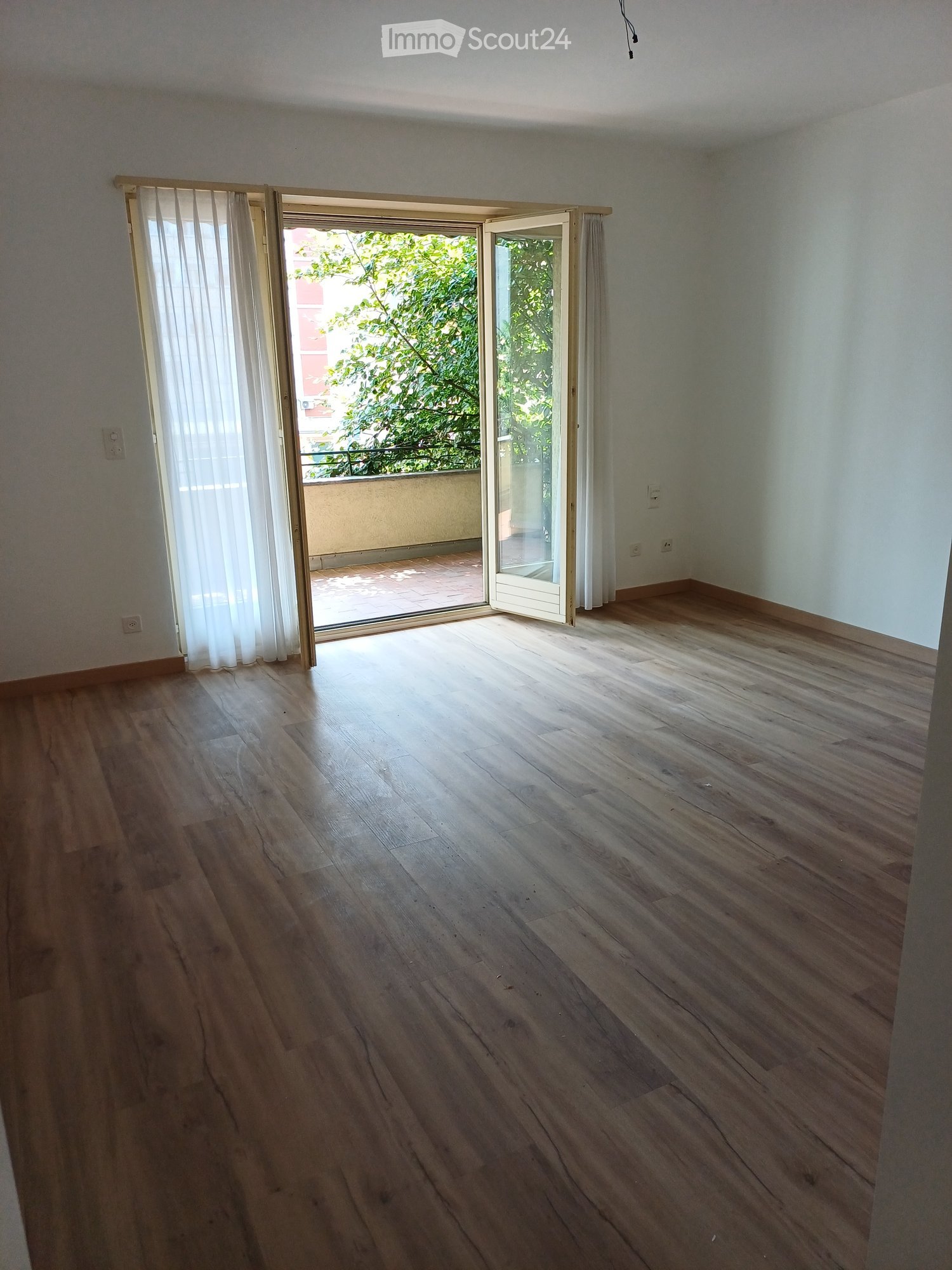 Empty room with wooden flooring, white walls, and large sliding glass doors leading to a balcony with outdoor space and greenery visible.