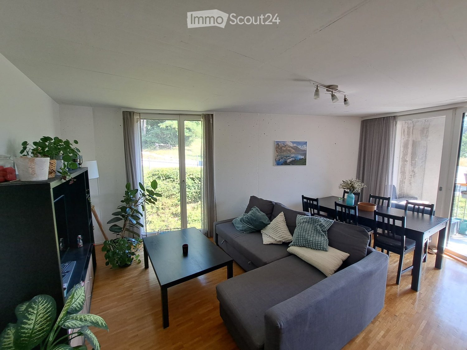 Living room with a grey sectional sofa, coffee table, dining table with chairs, and several potted plants. Large windows and sliding glass doors lead to an outdoor area.