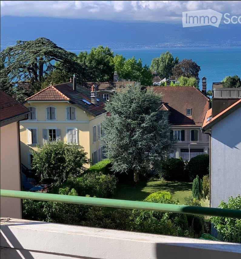 View from a balcony or terrace showing a garden area with trees and shrubs, overlooking a body of water and distant mountains.