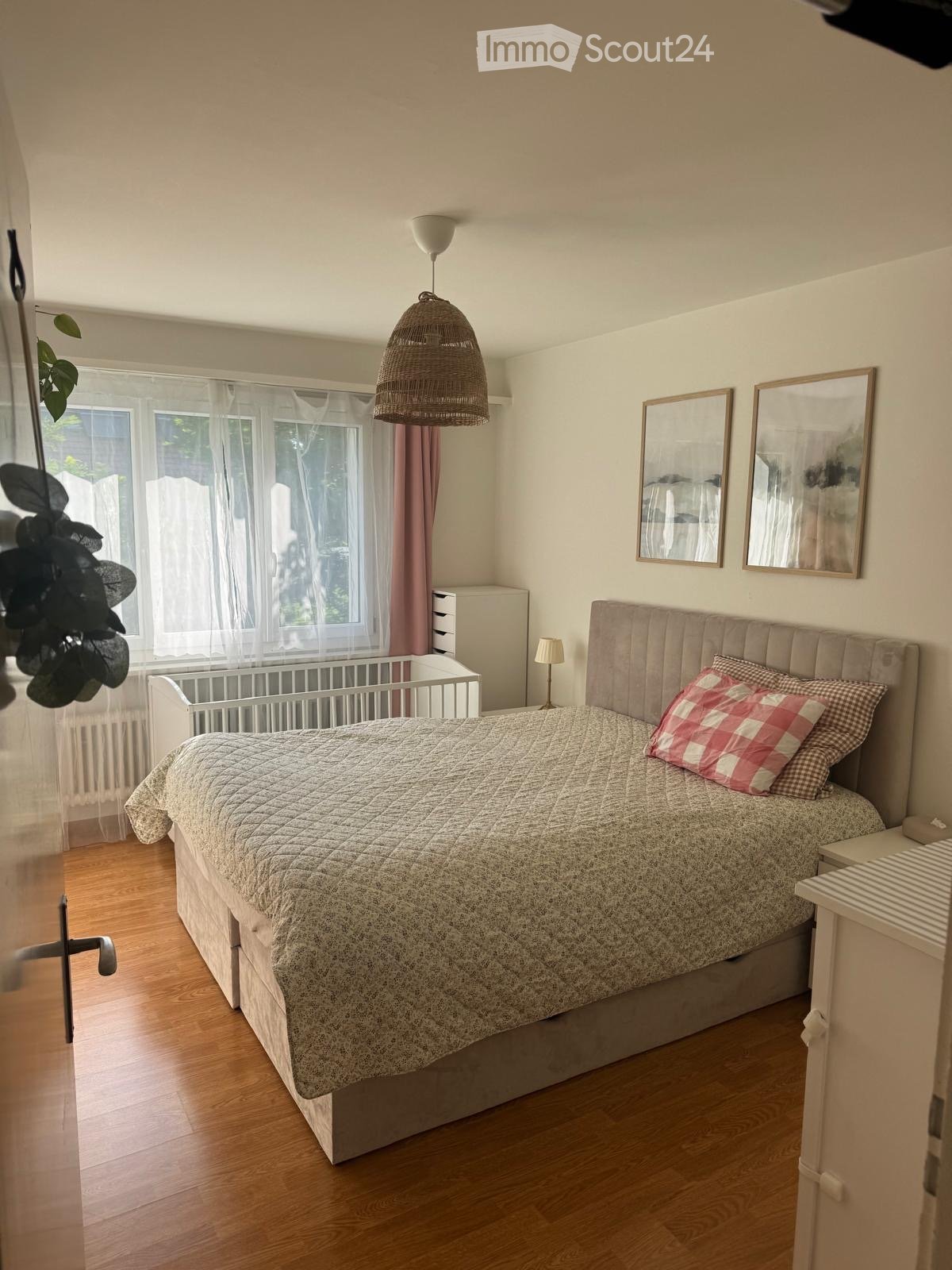 Cozy bedroom with wooden flooring, large window with white curtains, a double bed with a patterned quilt and pillows, a nightstand with a lamp, two wall-mounted mirrors, a wicker pendant light, a small plant, and a baby crib near the window.