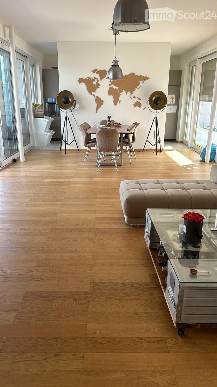 Open-plan living and dining area with wooden flooring, a large sofa, a coffee table, a dining table with chairs, pendant lights, and floor lamps.