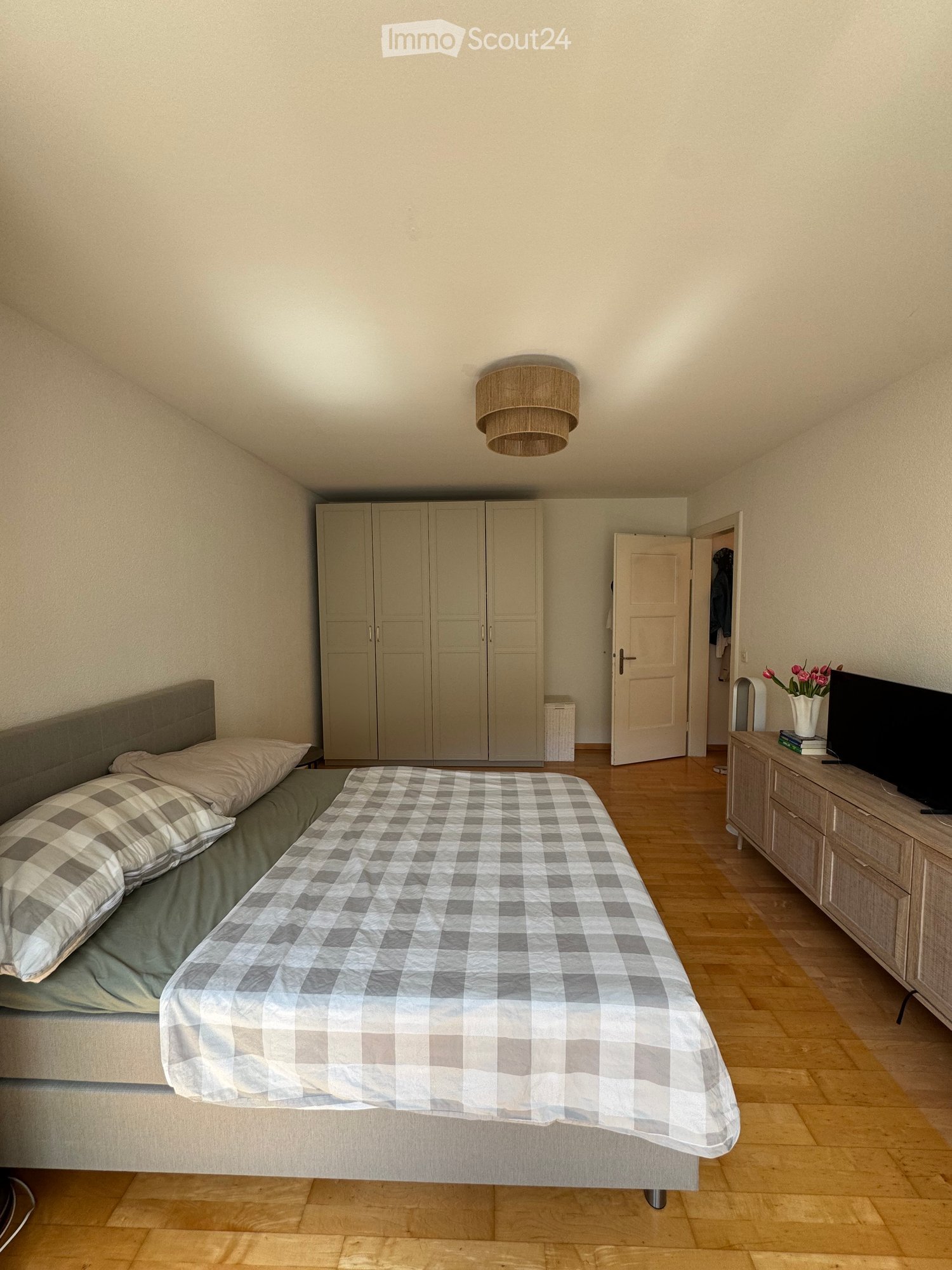 A well-lit bedroom with a double bed, checkered bedspread, pillows, a wardrobe, a nightstand with a TV, and a vase with flowers on the nightstand.