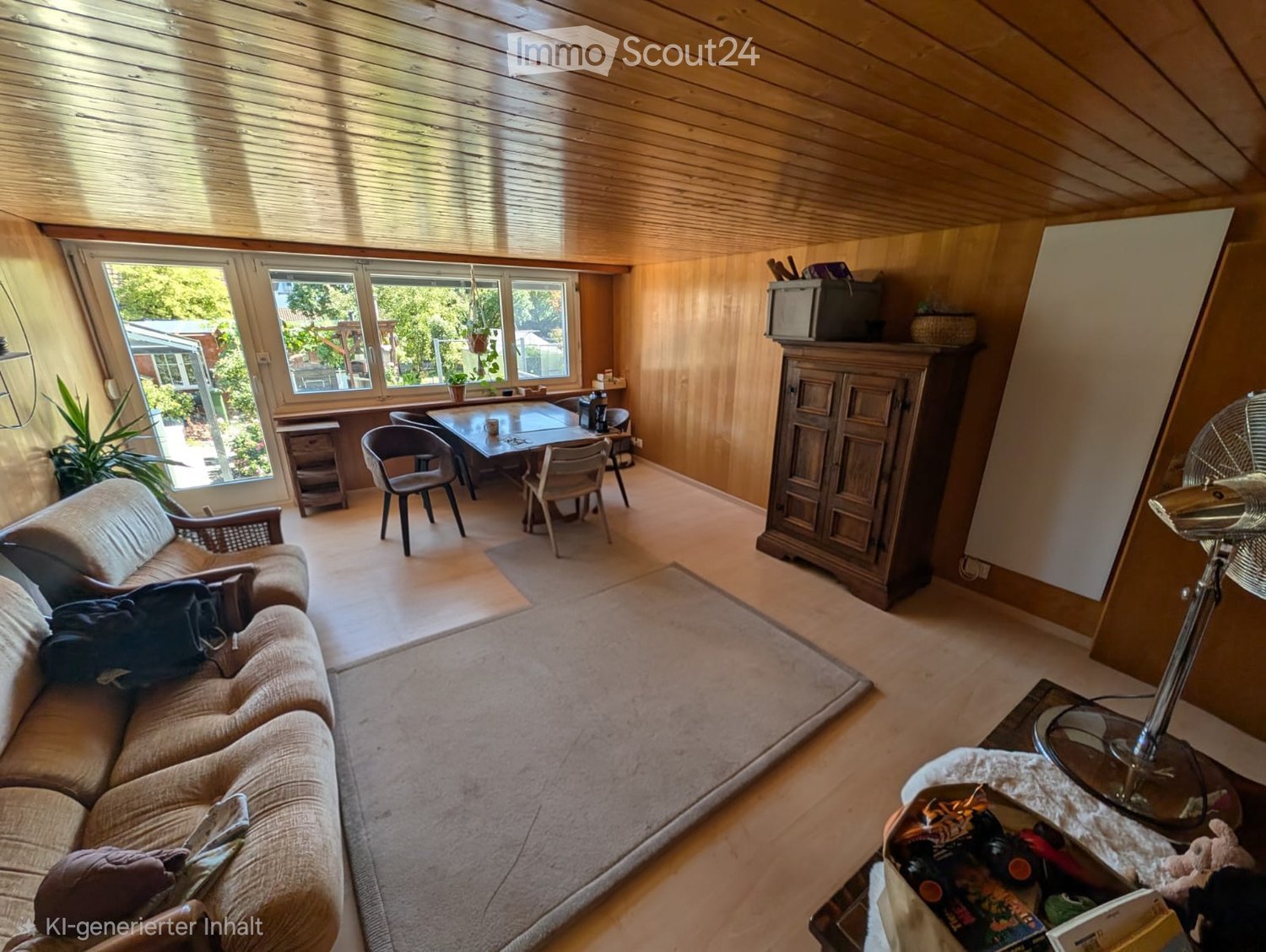 A cozy living room with wooden walls and ceiling, large windows, a sofa, a coffee table, a dining table with chairs, a cabinet, and a fan.
