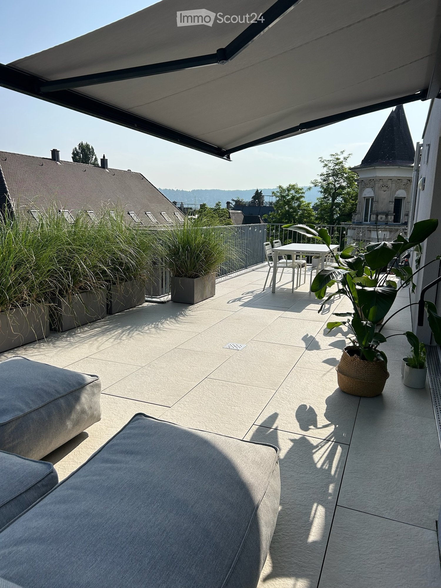 A modern balcony with a tiled floor, a grey sofa, a small table with chairs, and several potted plants, offering a scenic view of the city and surrounding landscape.