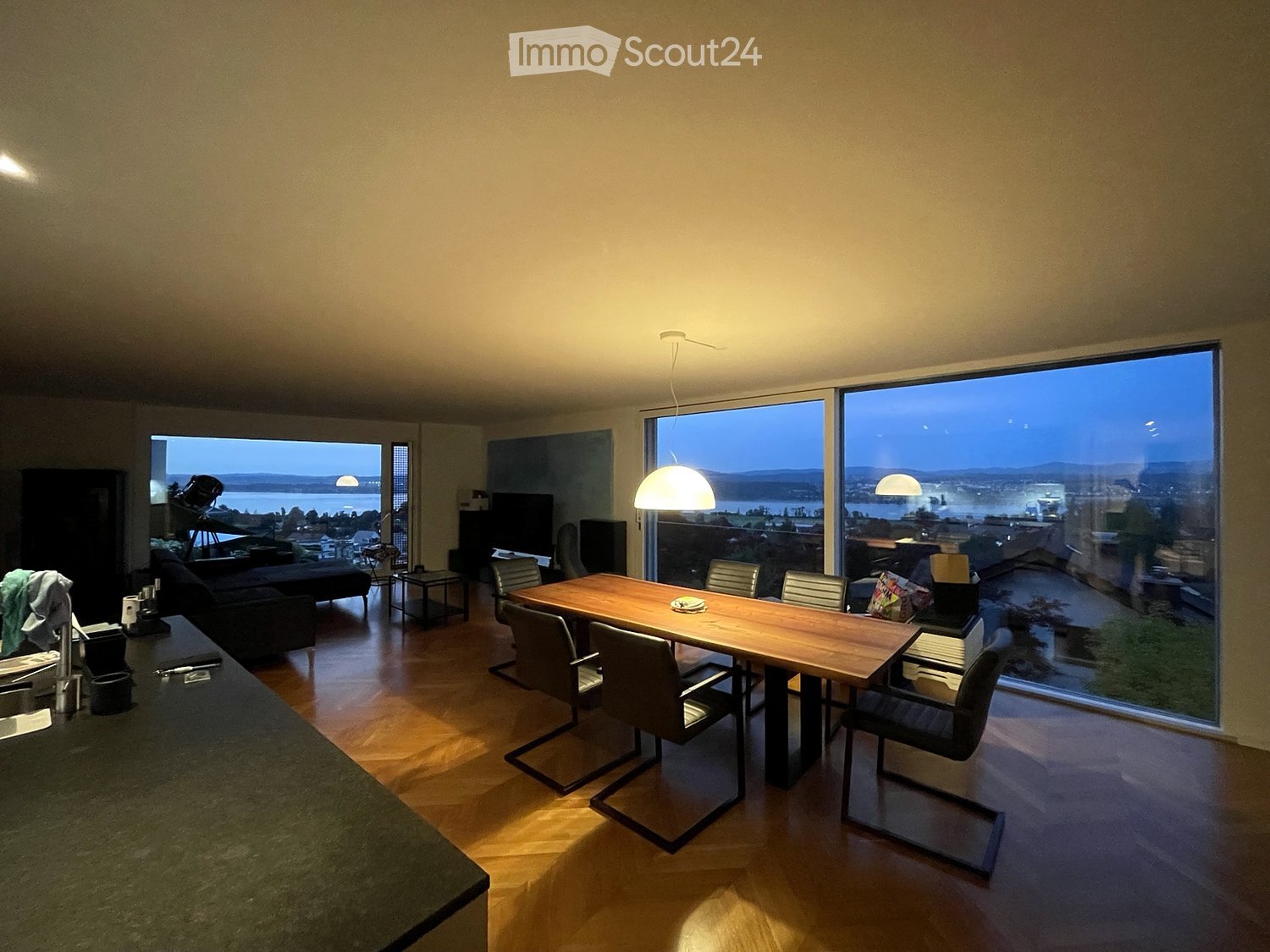 Open-plan living and dining area with large windows offering a scenic view, featuring a sofa, dining table with chairs, and a television.