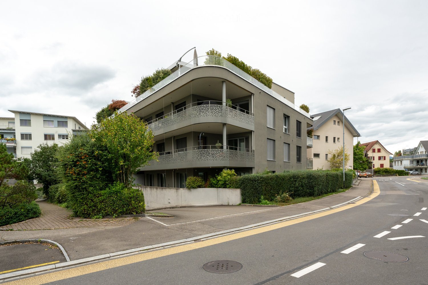 A multi-story building with a green terrace, a balcony with a lacy design, and parking in front.