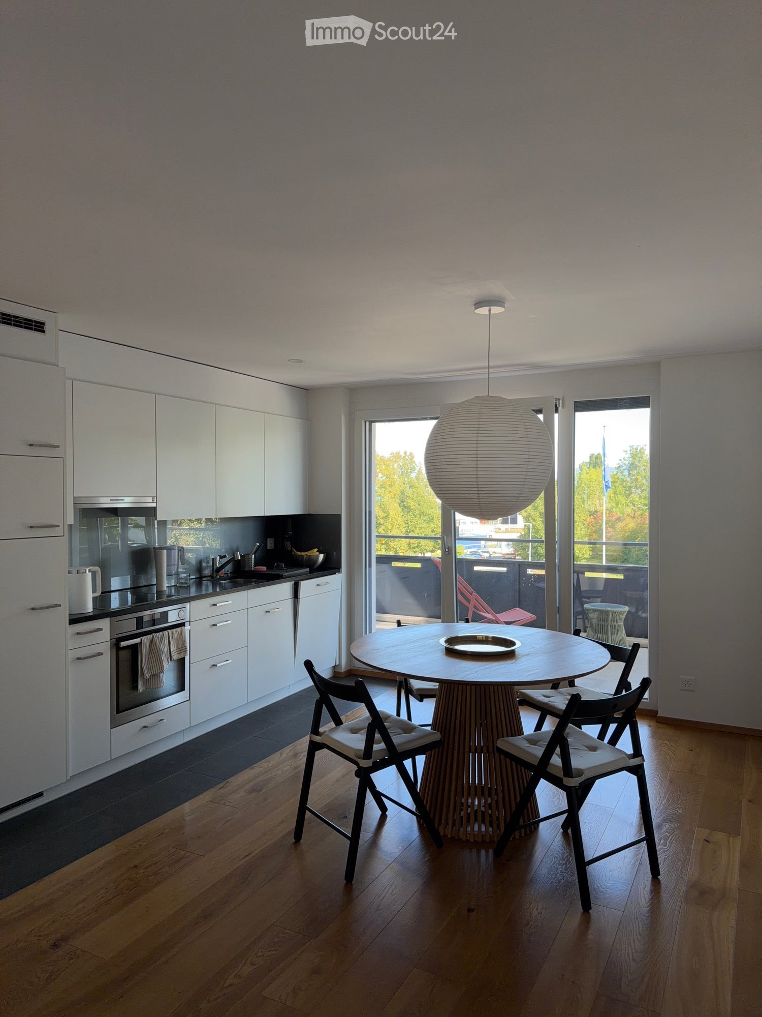 Modern kitchen with white cabinets, stainless steel appliances, a round wooden dining table with four chairs, and a hanging spherical lamp.