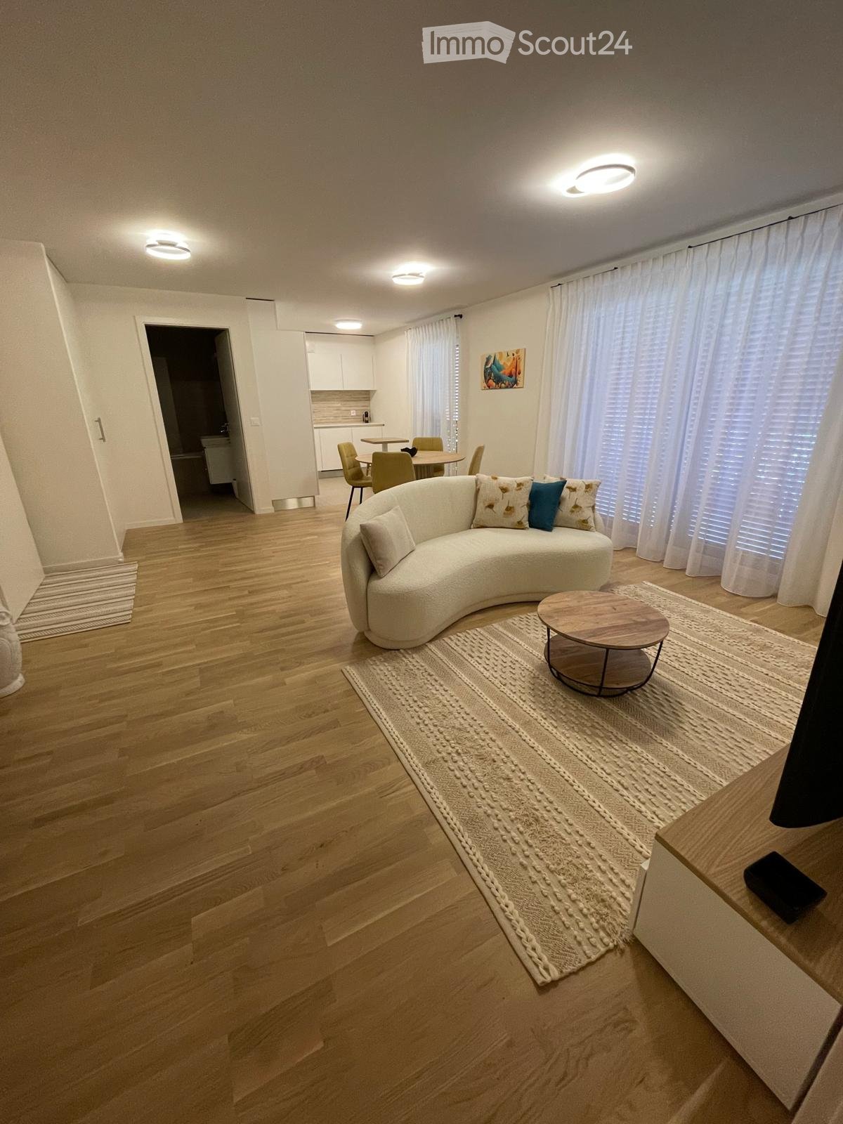 Modern living room with a curved white sofa, coffee table, dining area with chairs, large windows with curtains, and artwork on the wall.