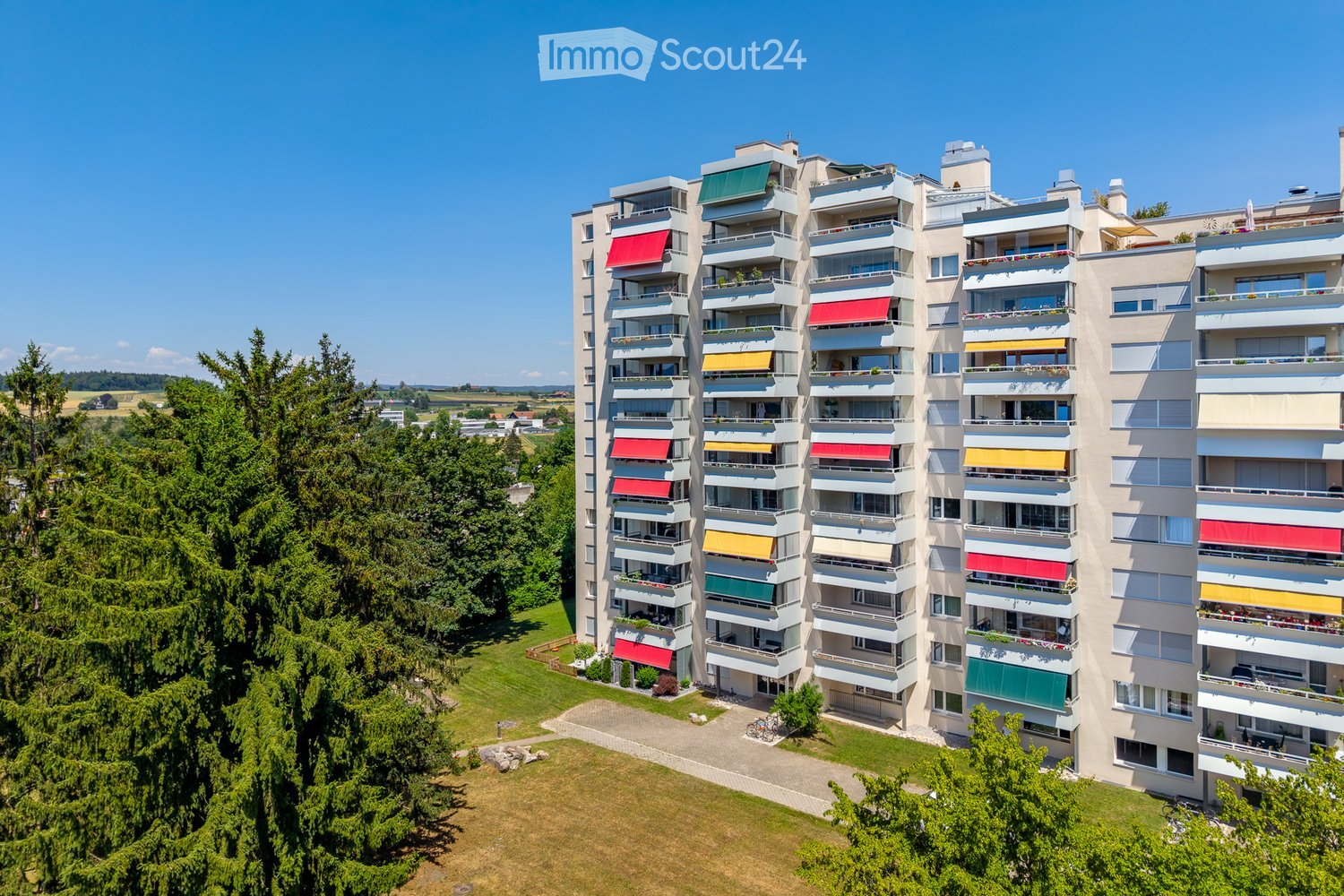 A modern multi-story apartment building with balconies, colorful awnings, and surrounding greenery.